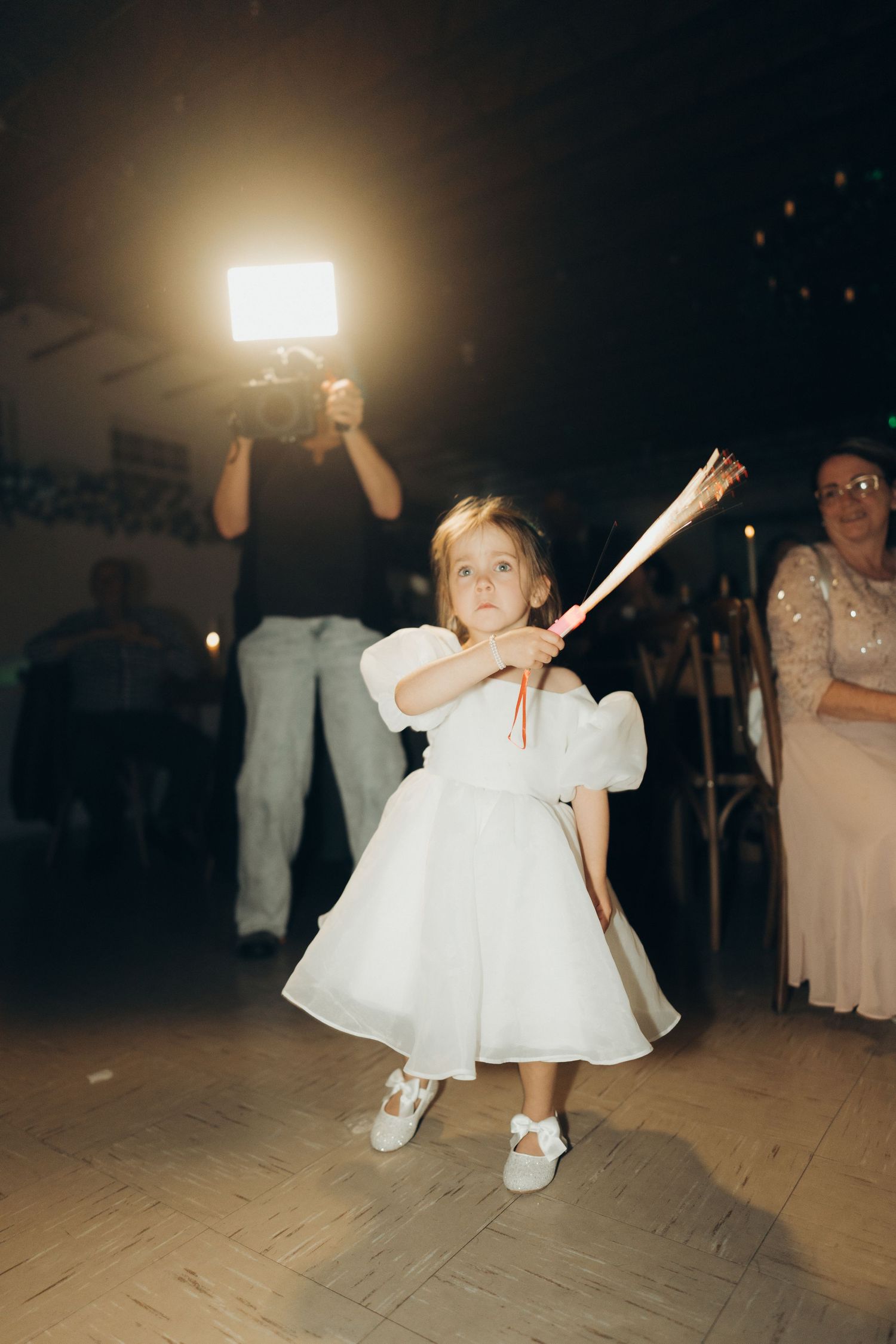 A young child in a white party dress dances energetically on a wooden dance floor at an evening celebration.