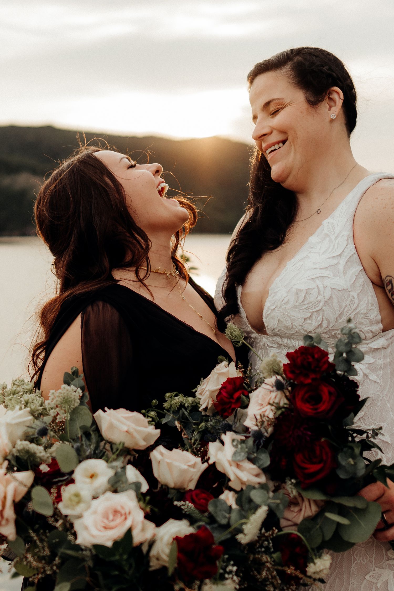 Two brides share a joyful laugh while holding beautiful bouquets of red and white roses at sunset.