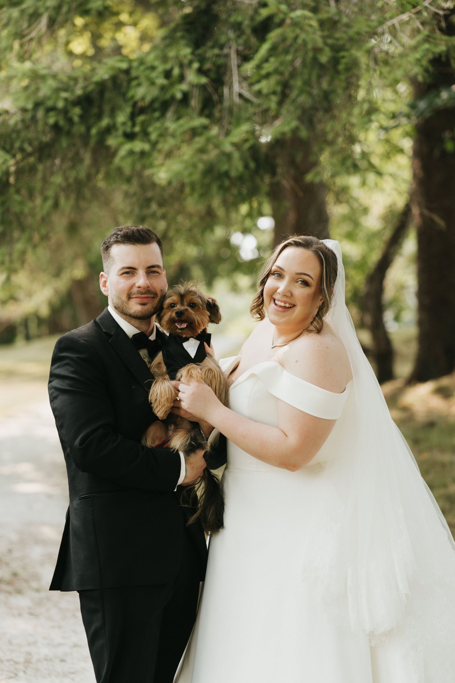 Newlyweds in formal attire play with their small brown dog in a wooded outdoor setting.
