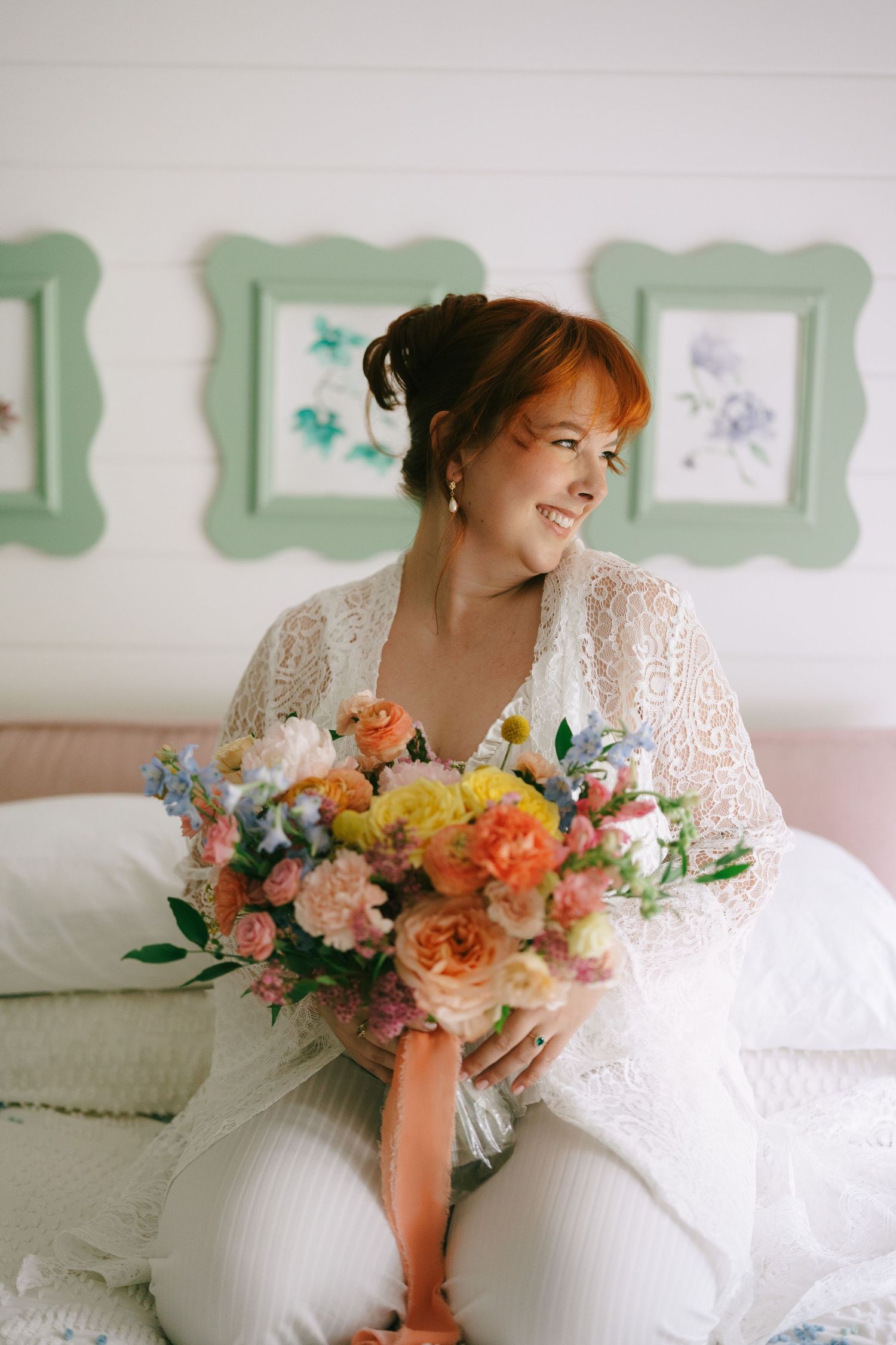 Smiling bride in lace robe holding colorful floral bouquet with pink ribbon, sitting on bed with green framed art.