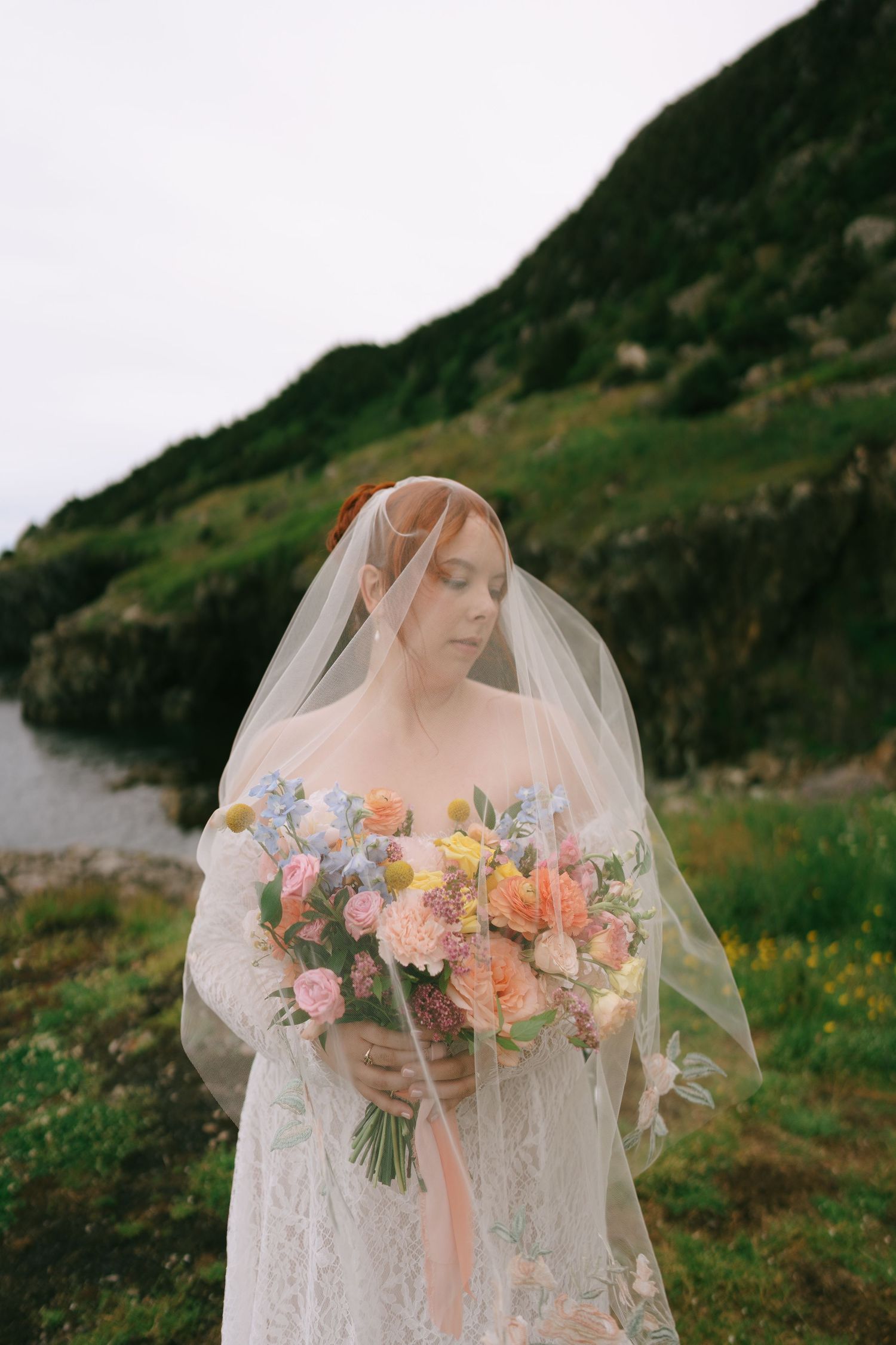 Bride in lace gown and veil holds colorful bouquet of pastel flowers outdoors near rocky green hillside.