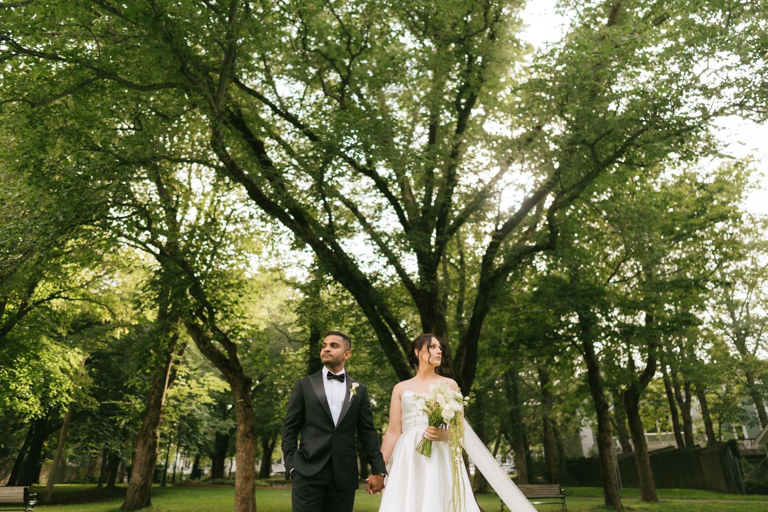 Bride and groom holding hands in a lush green park surrounded by tall trees on their wedding day.
