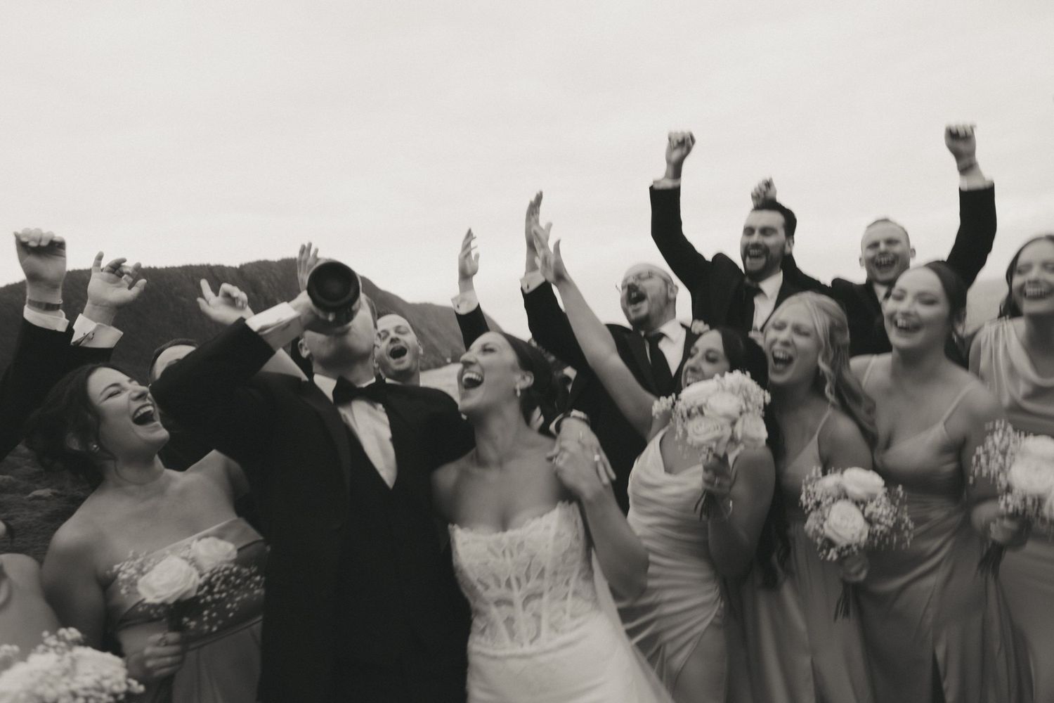 Black and white wedding party photo with bride, groom, and guests cheering joyfully with arms raised.