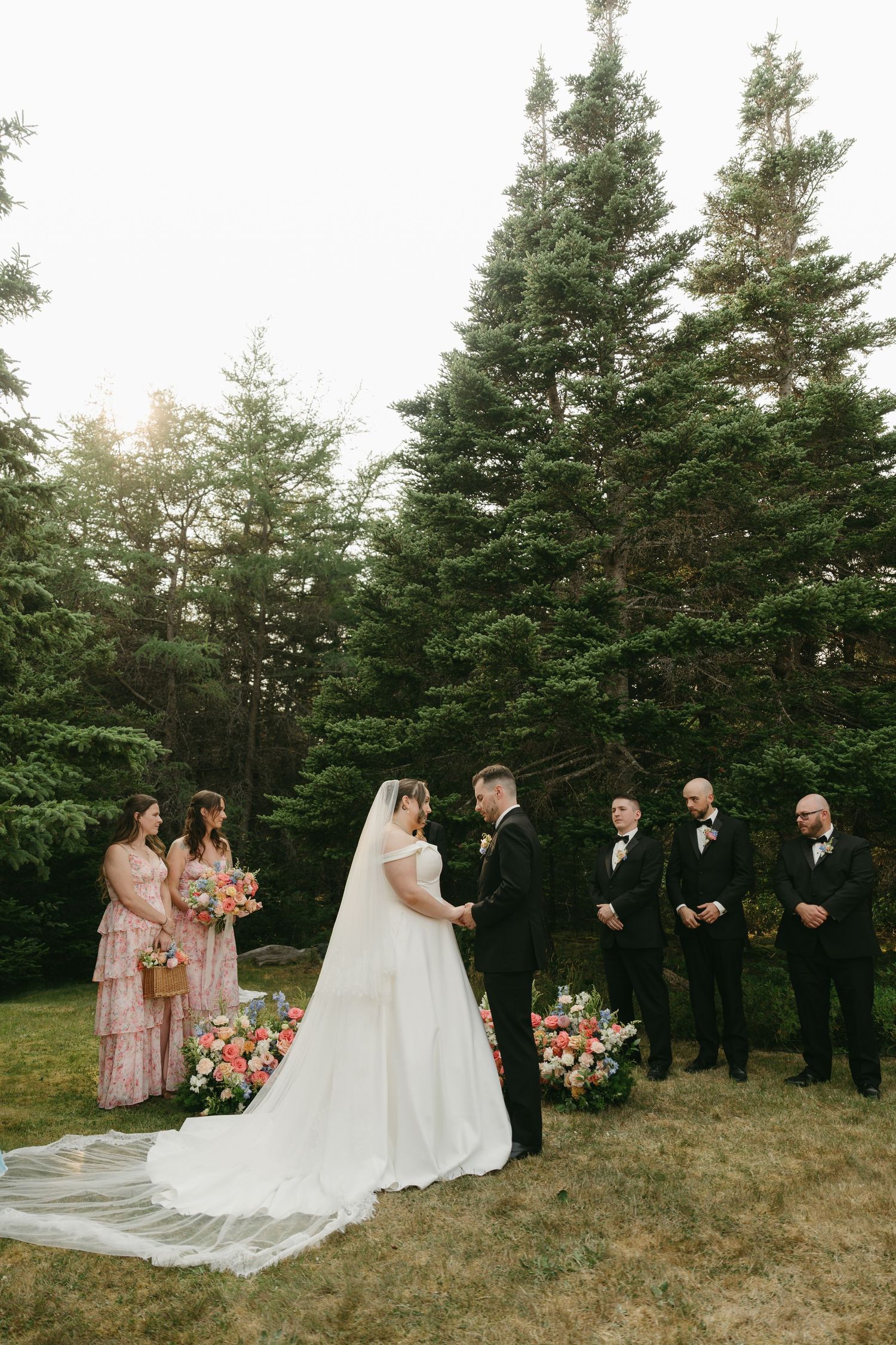 Forest wedding ceremony with bridal party in formal attire standing before tall pine trees.