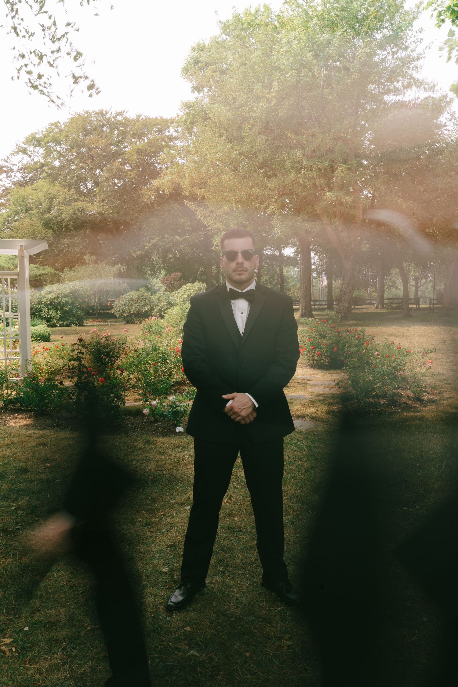 Groomsmen in black tuxedos pose outdoors in natural sunlight.
