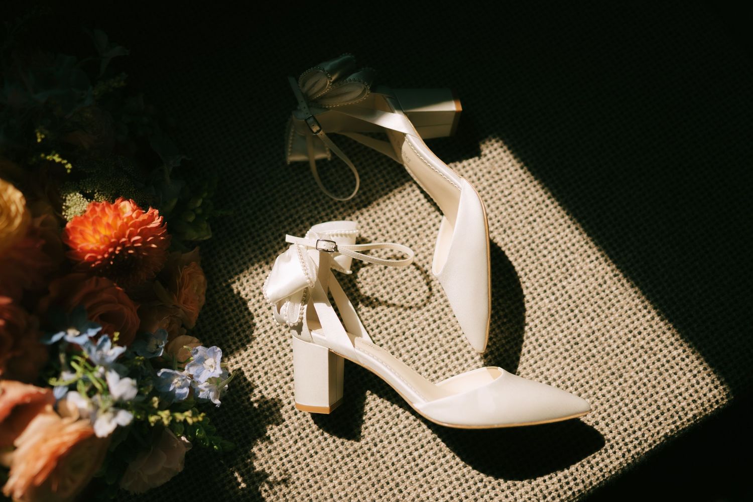 Wedding accessories including shoes and flowers arranged on a textured surface in dramatic lighting.