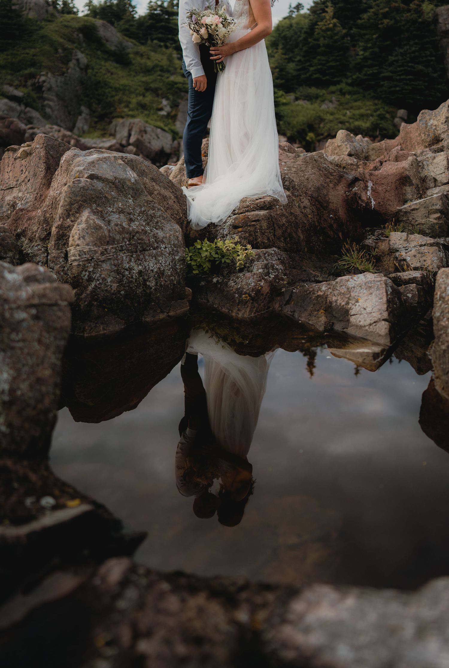 Wedding couple's reflection captured in tide pool among coastal rocks.