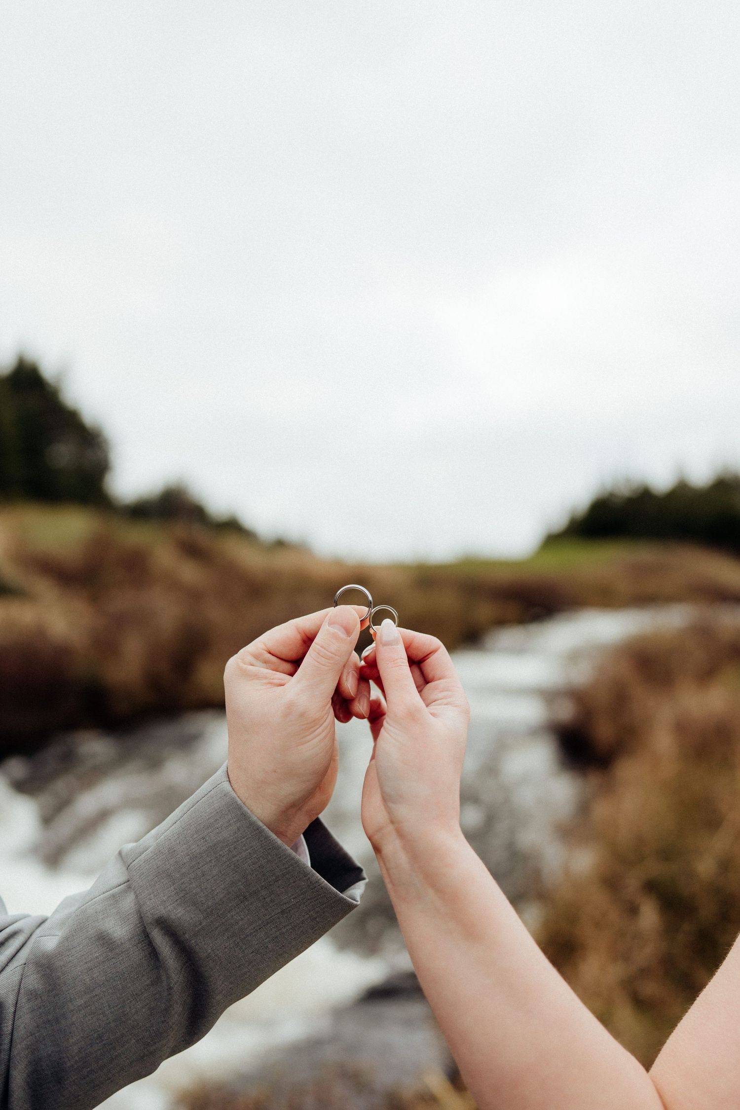 Close-up of hands holding wedding rings against a coastal background.