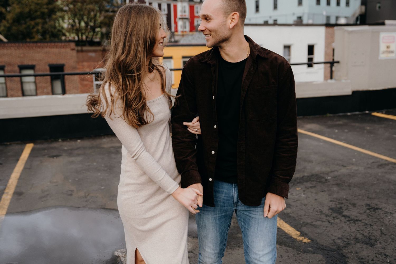 A couple in casual attire embraces lovingly in a parking lot with city buildings in the background.