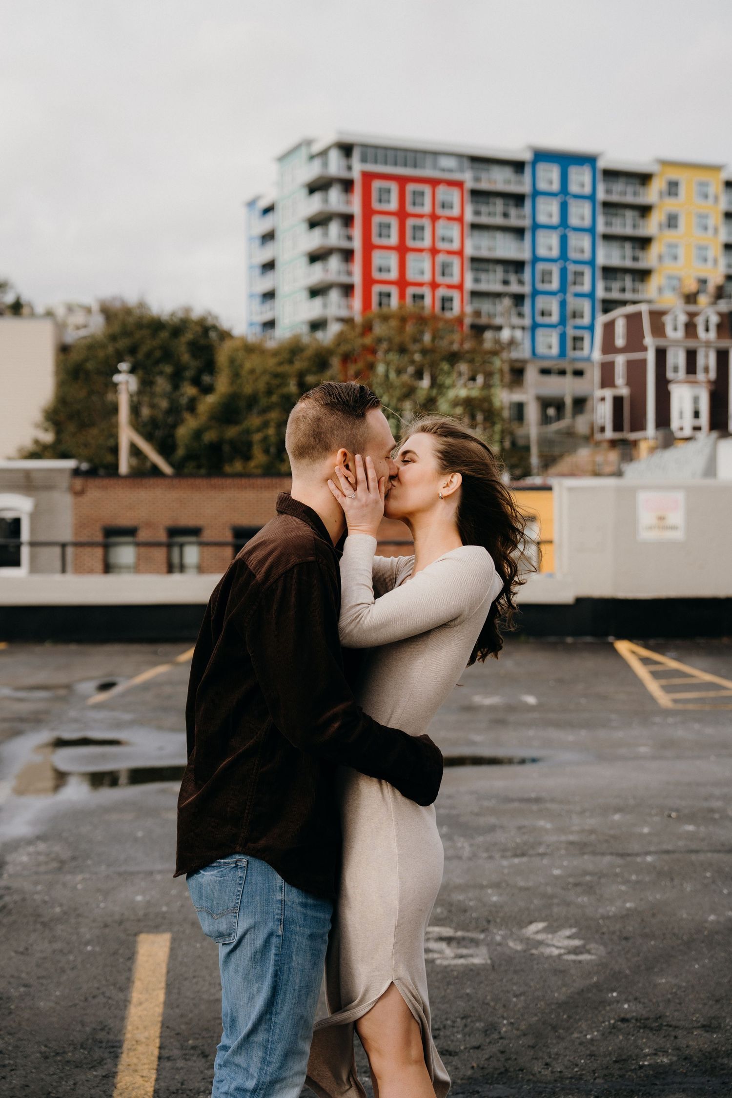 A romantic couple embraces in an empty parking lot with modern apartments behind them.