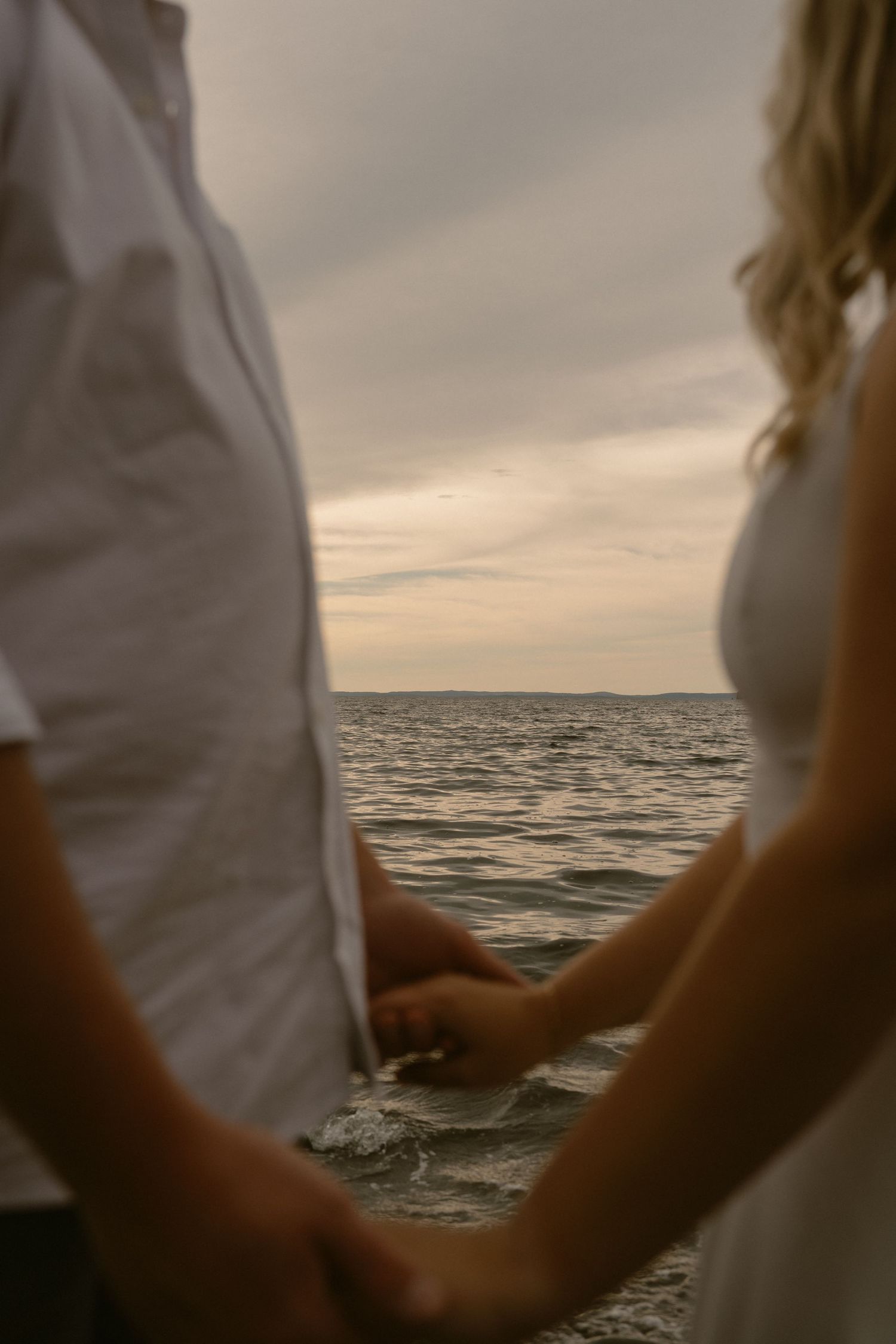 A couple holds hands against a moody sunset seascape with waves rolling in the background.