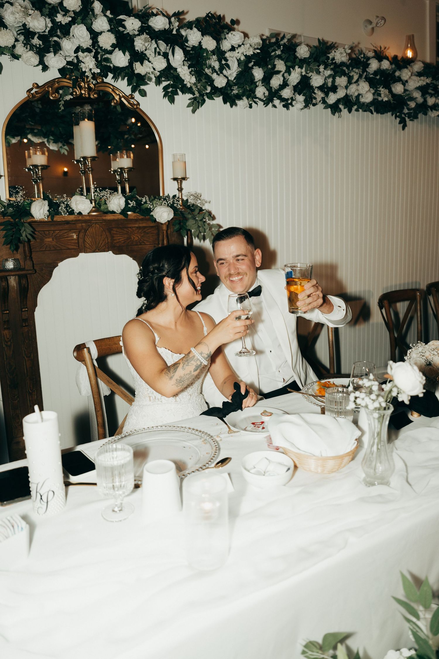 A couple toasts at their wedding reception table decorated with white linens and floral garlands.