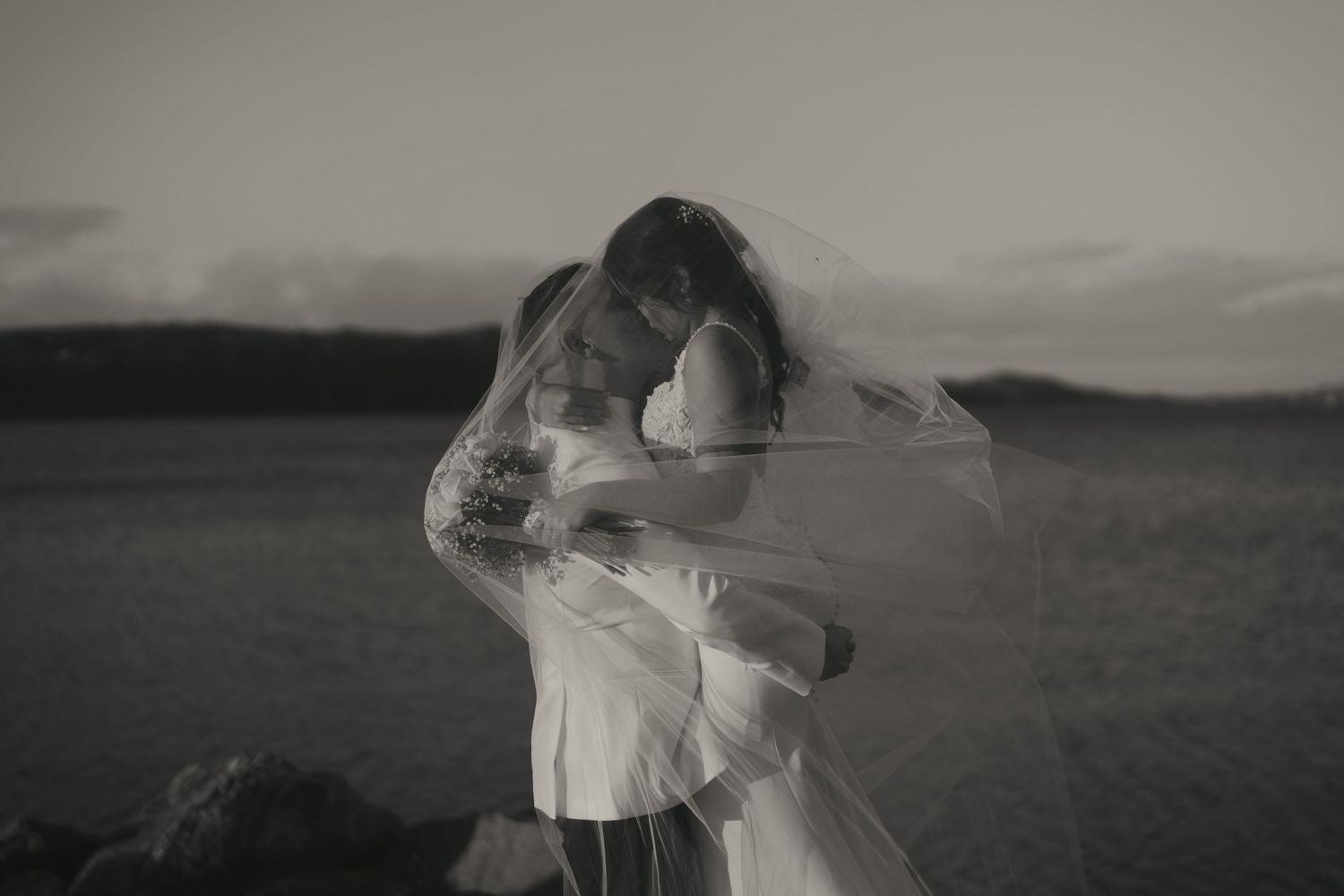 A romantic couple embraces while standing in a field at sunset creating a silhouette against the dramatic sky.