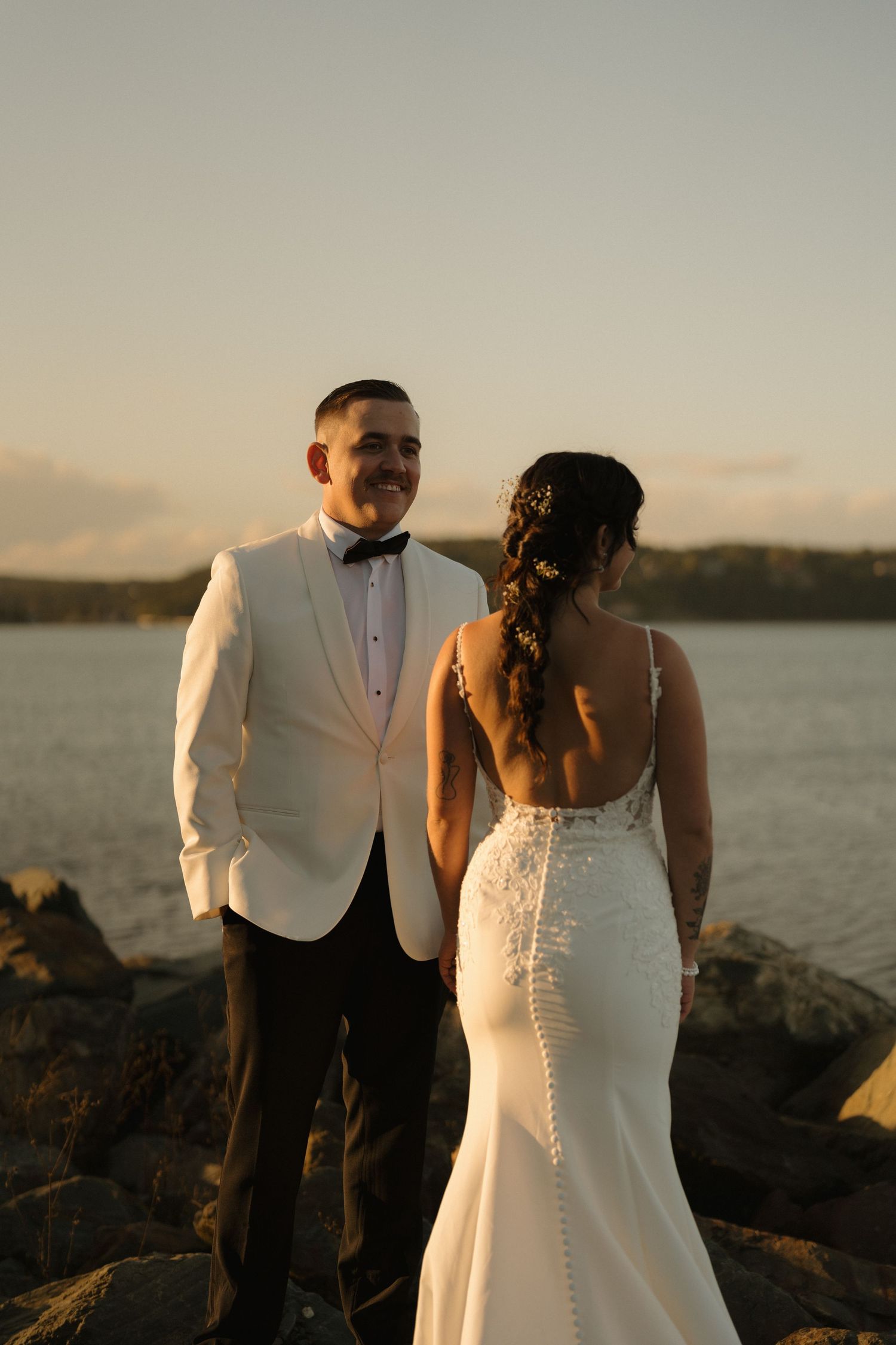 A couple in wedding attire stands together on rocky coastal terrain at sunset with a waterfront backdrop.