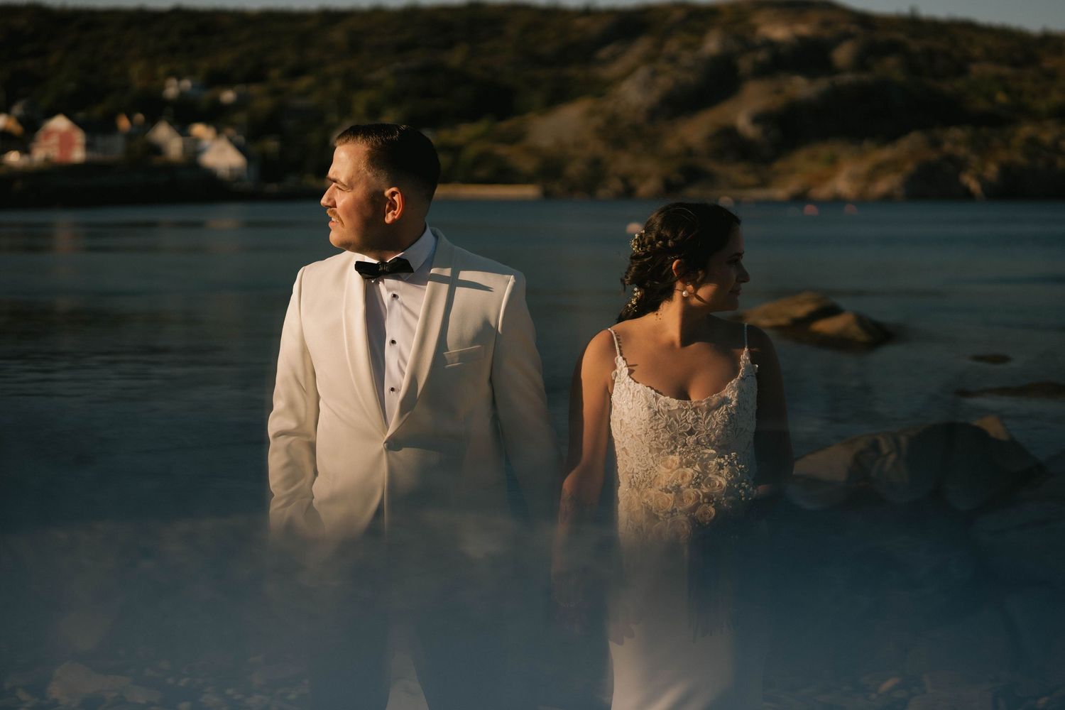 A romantic couple in wedding attire stands together against a scenic lakeside backdrop at sunset.