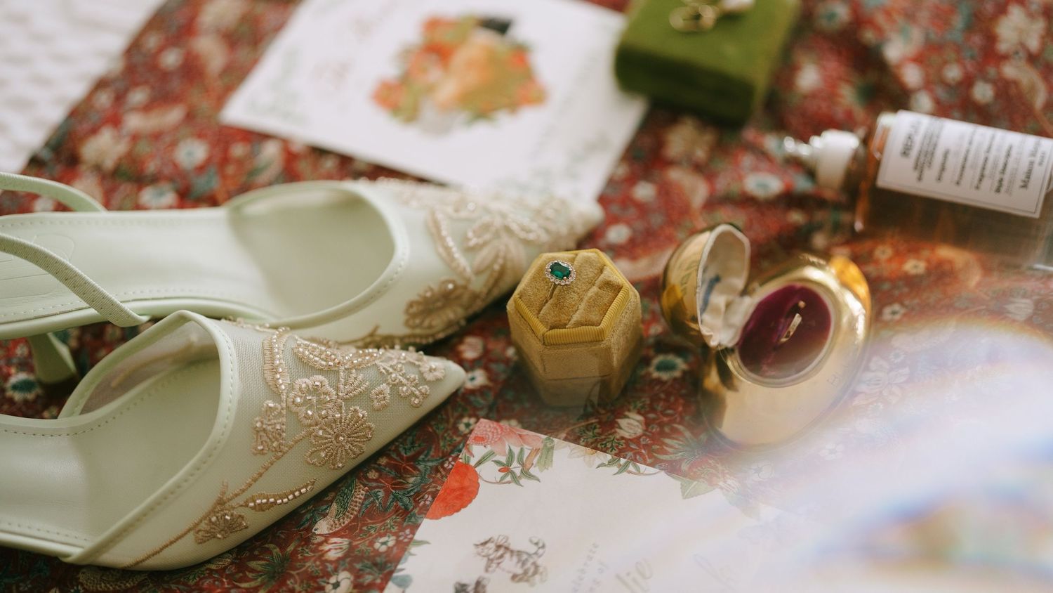 Elegant bridal flat shoes with floral embroidery beside gold ring boxes and jewelry on a floral fabric backdrop.