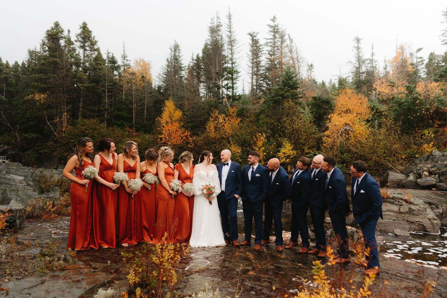 Wedding party in red dresses and navy suits poses on rocky terrain with autumn trees and foliage in background.