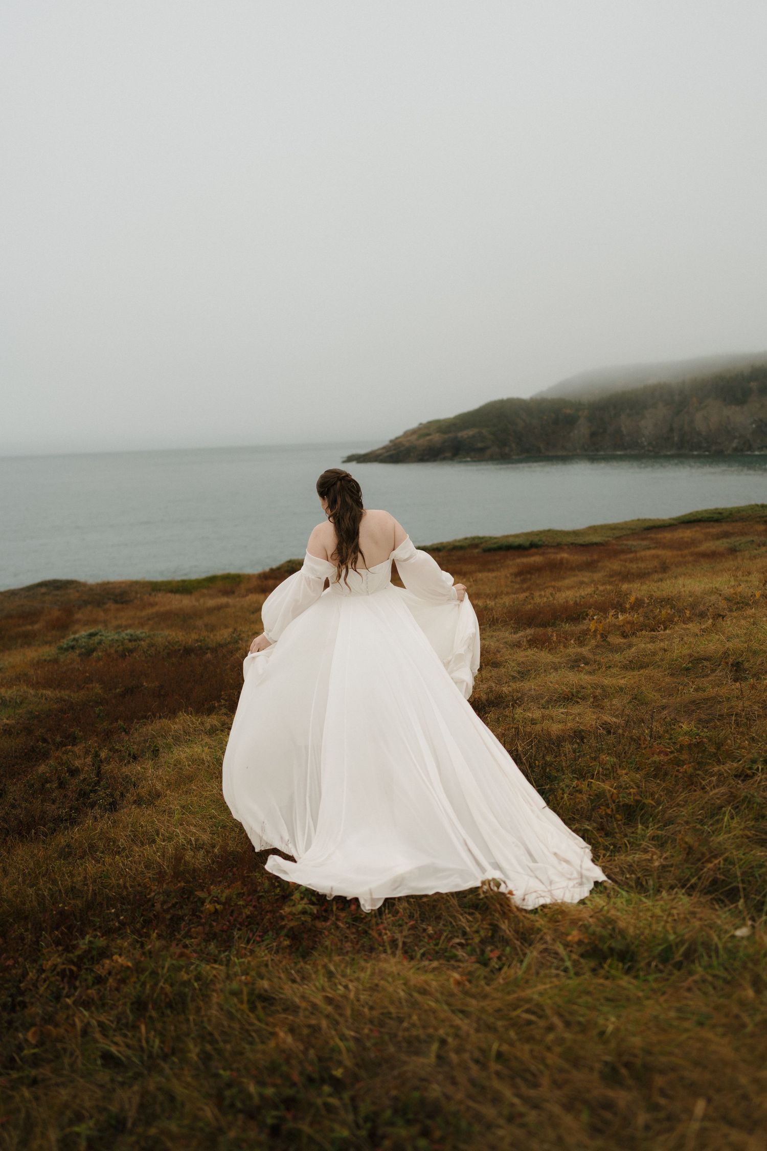 A person in a flowing white dress stands on a misty coastal cliff overlooking the ocean on a moody day.