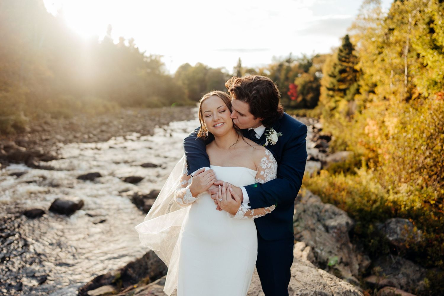 Newlyweds share an intimate embrace by a rocky riverbank during autumn.