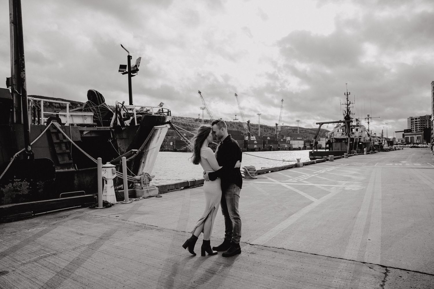 Black and white image of couple dancing on waterfront with boats and port facilities visible.