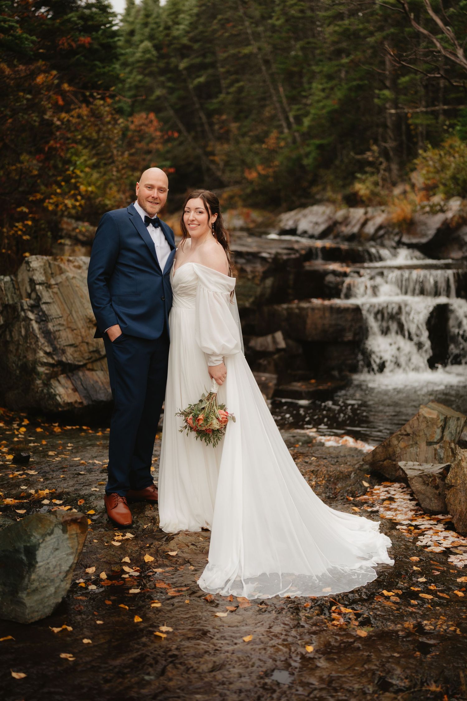 A couple poses in wedding attire by a scenic waterfall surrounded by autumn foliage and rocky terrain.