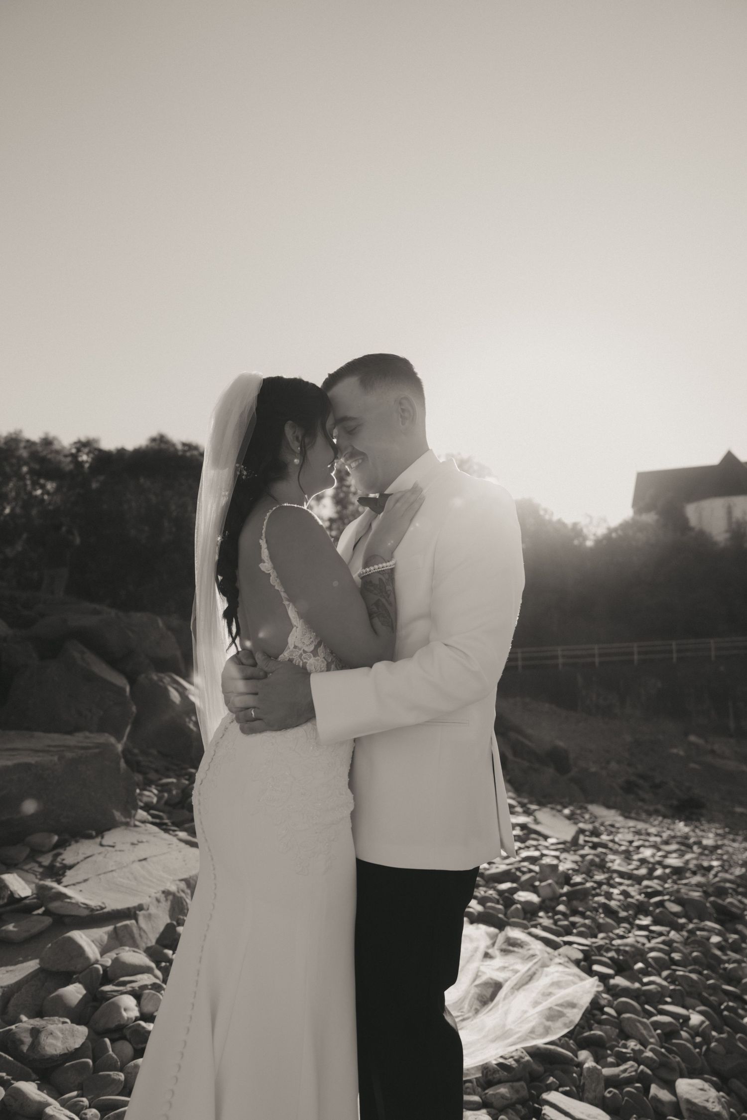 A romantic black and white photo series showing a couple embracing on a rocky beach at sunset.