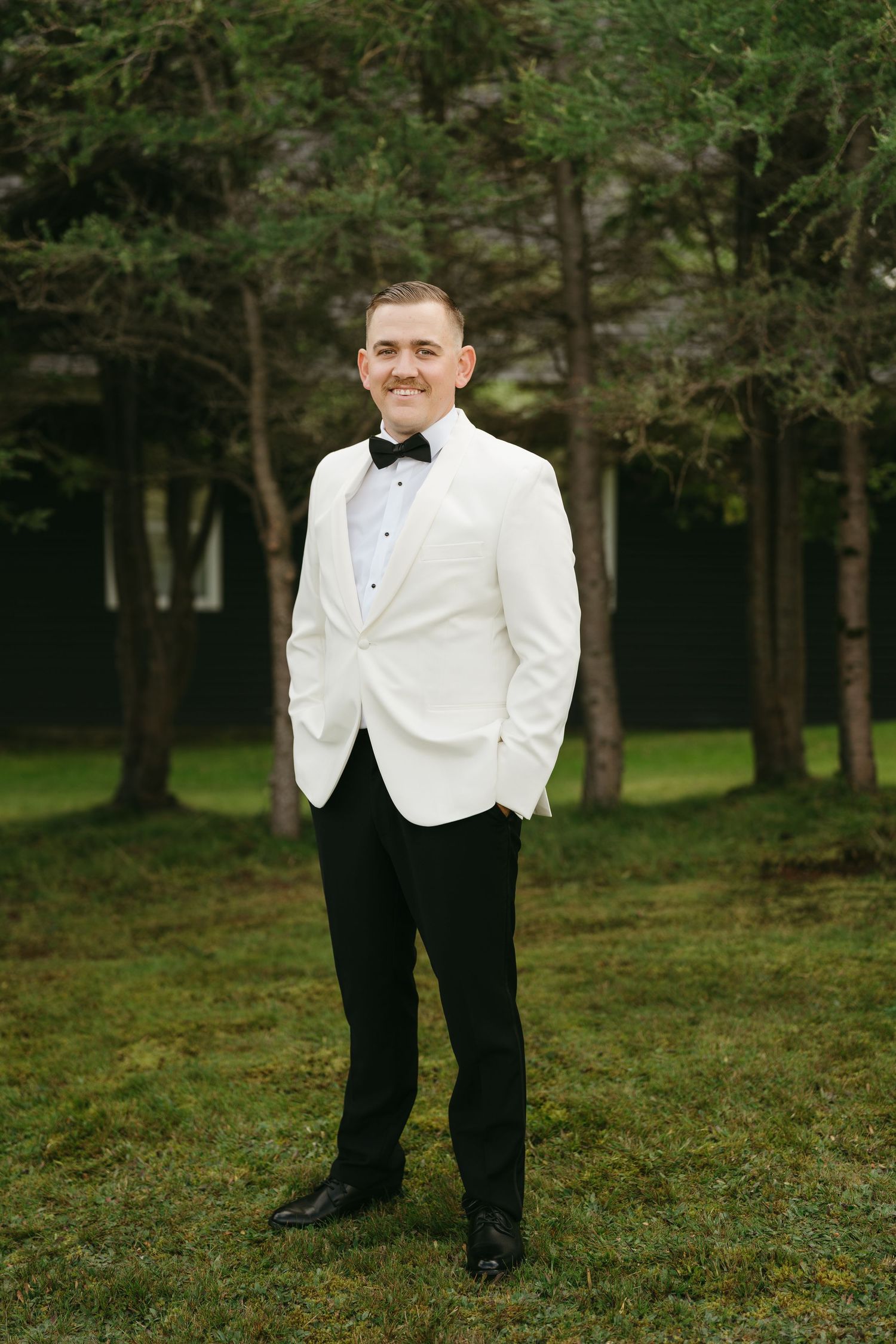 A person in a white dinner jacket and black bow tie stands in a wooded outdoor setting.