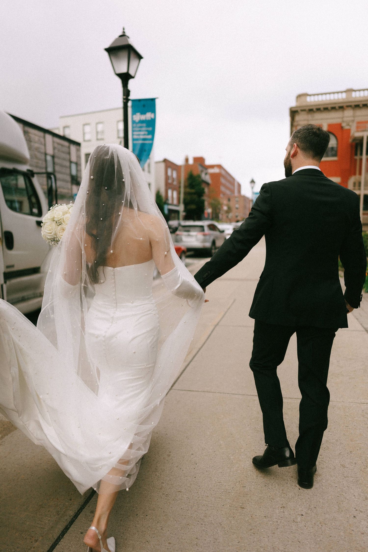 Bride and groom walking hand in hand down a city sidewalk, bride wearing a flowing white gown and veil with bouquet.