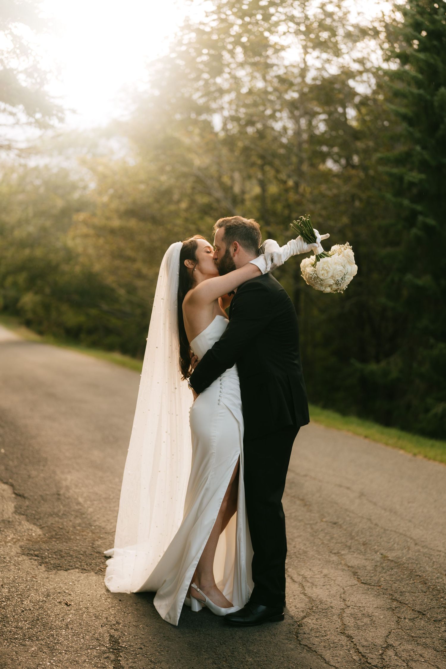 Bride and groom share a romantic kiss on a sunlit road, bride holding white bouquet in elegant gown with veil.