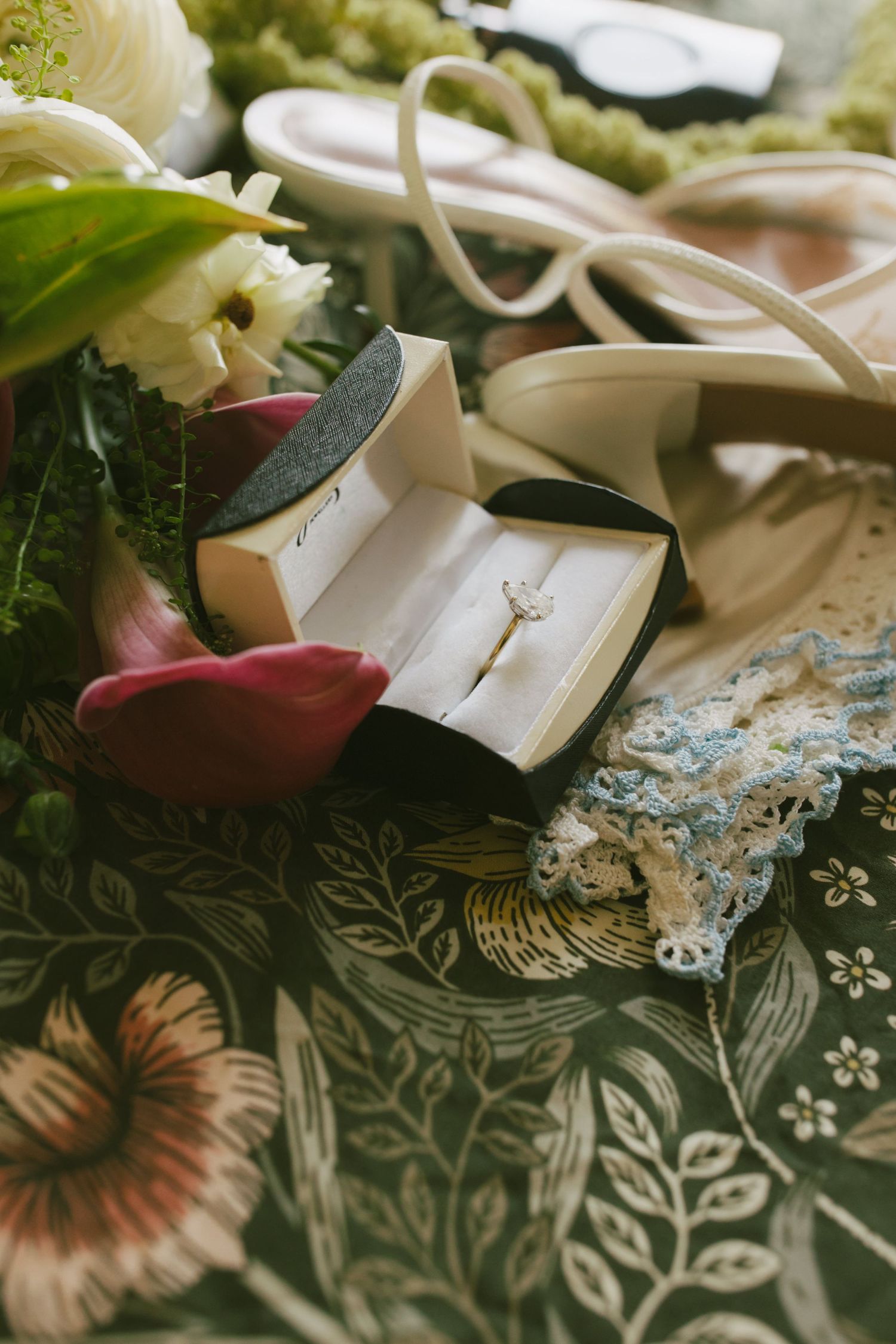 Diamond engagement ring in a black box beside bridal bouquet, heels, and lace garter on floral fabric.
