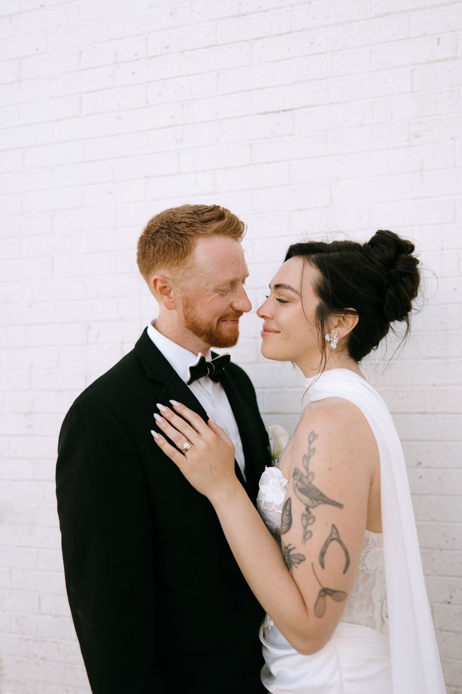 Bride and groom nearly kissing against white brick wall, bride showing diamond ring on grooms chest.
