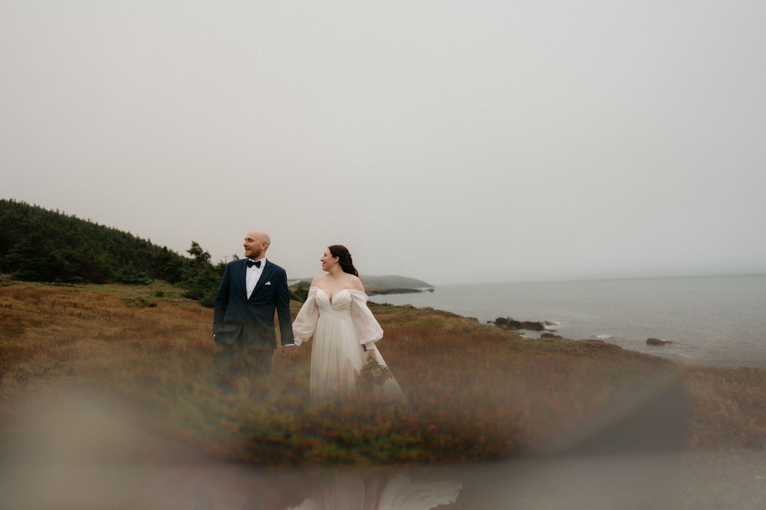 A couple in wedding attire walks along a misty coastal cliff with dramatic ocean views in the background.