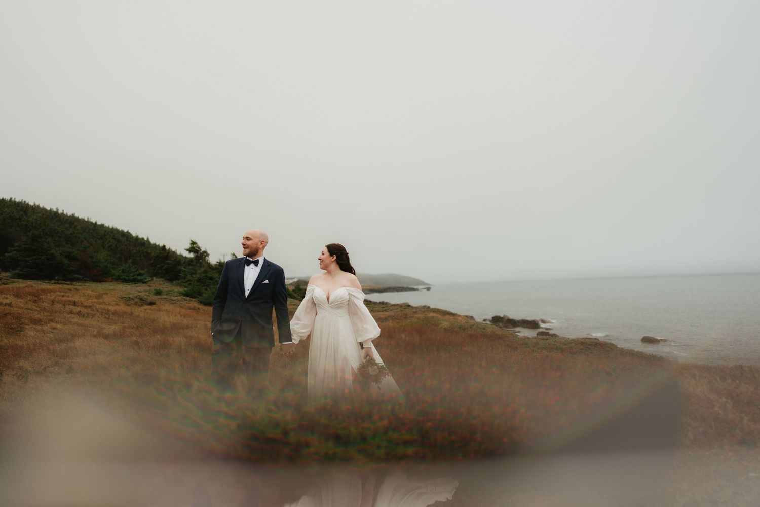 Two people walk along a misty coastal cliff path in wedding attire during an overcast day.