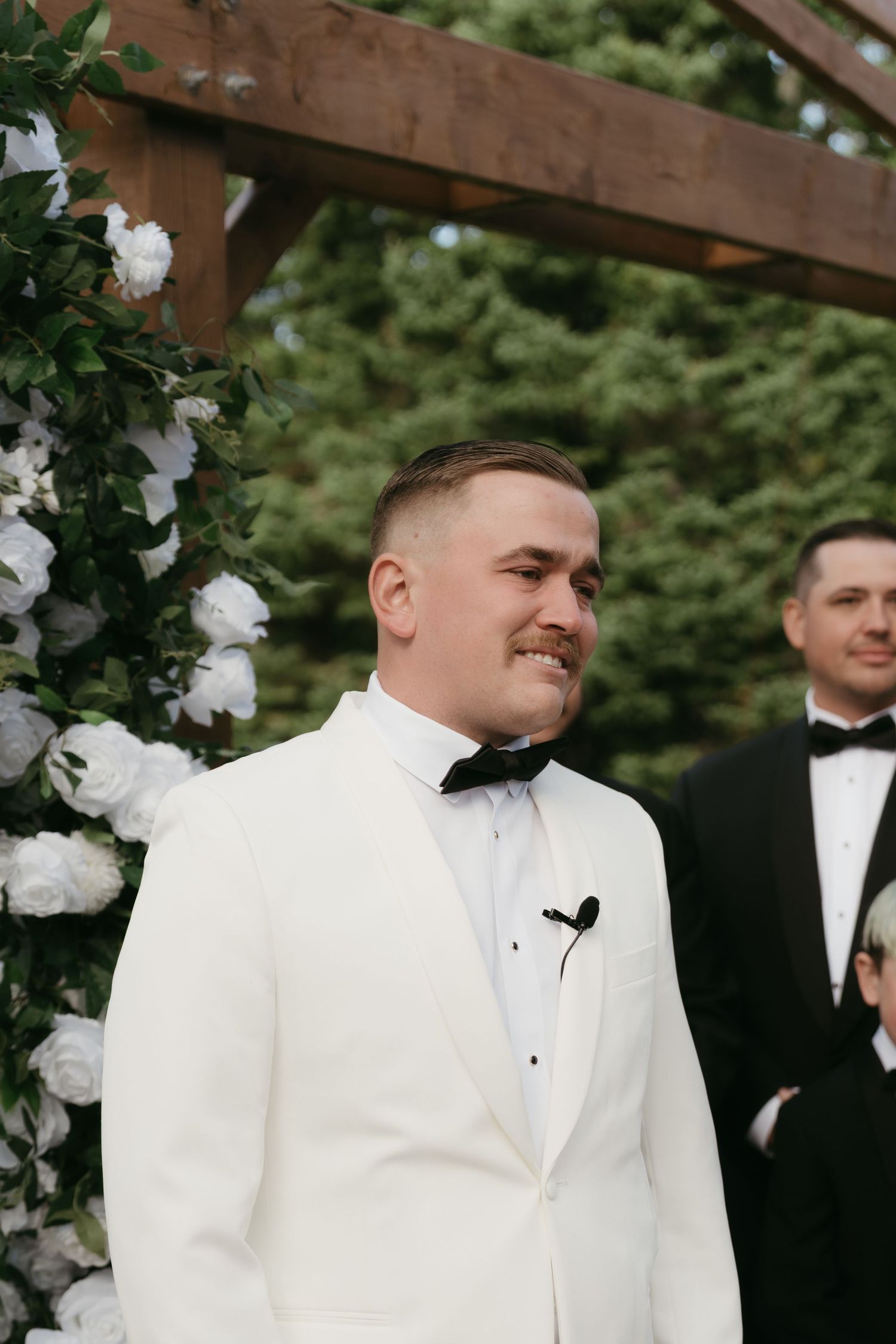 A groom in a white tuxedo jacket and black bow tie shows emotional reactions during an outdoor wedding ceremony.