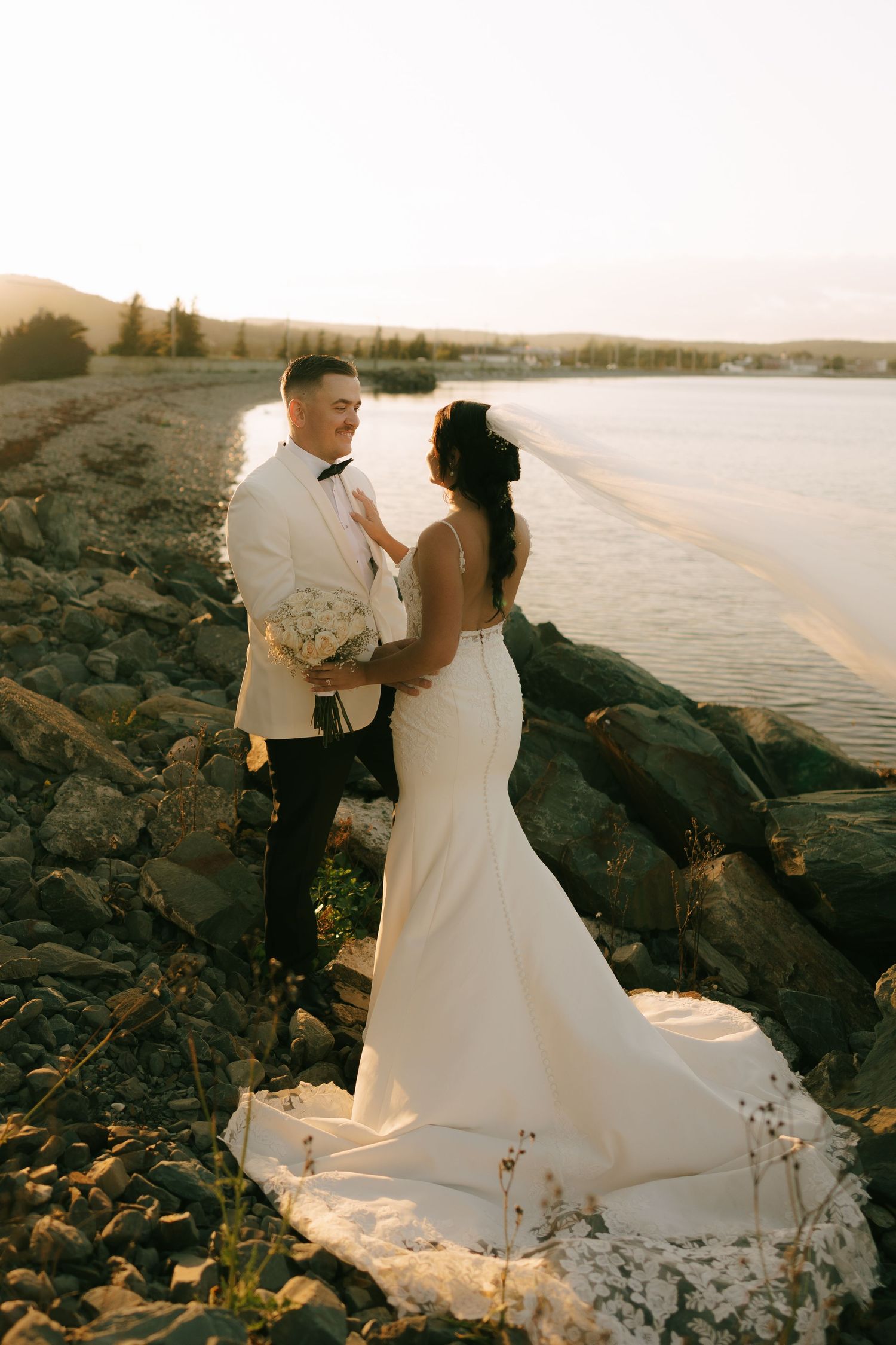 Newlyweds share intimate moments on a rocky beach at sunset with flowing white dress and ocean backdrop.