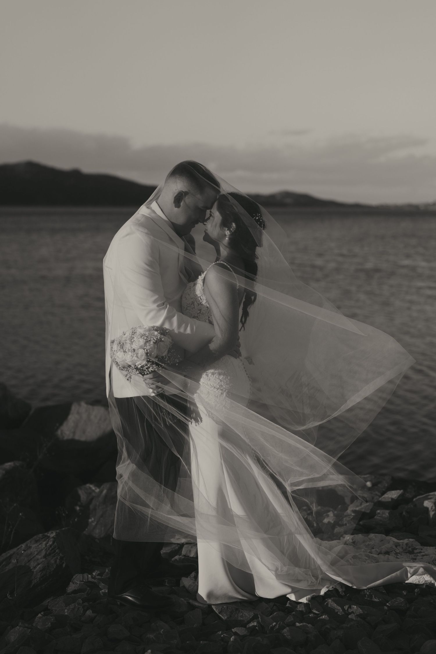 A romantic black and white wedding photo shows a couple embracing on a rocky shore with a flowing veil in the wind.