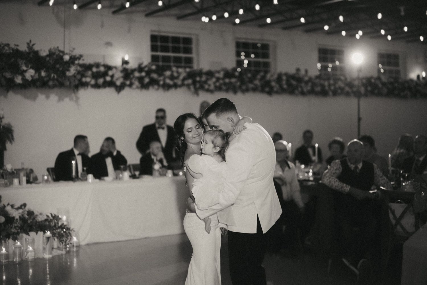 A romantic first dance at a wedding reception with twinkling lights and guests watching in the background.