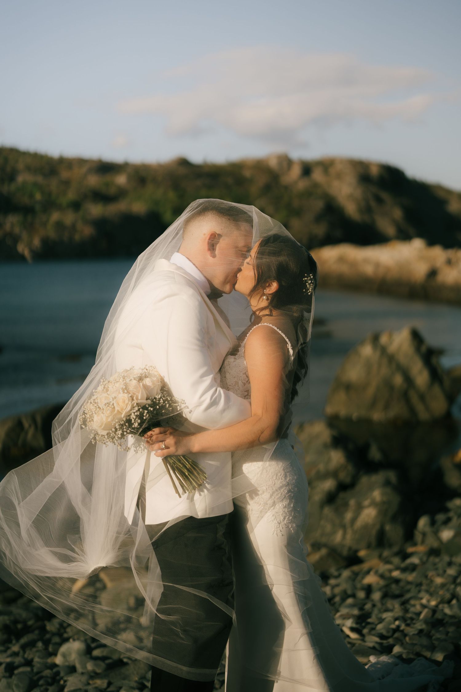 Newlywed couple shares romantic kisses on rocky beach at sunset with flowing veil and bouquet.