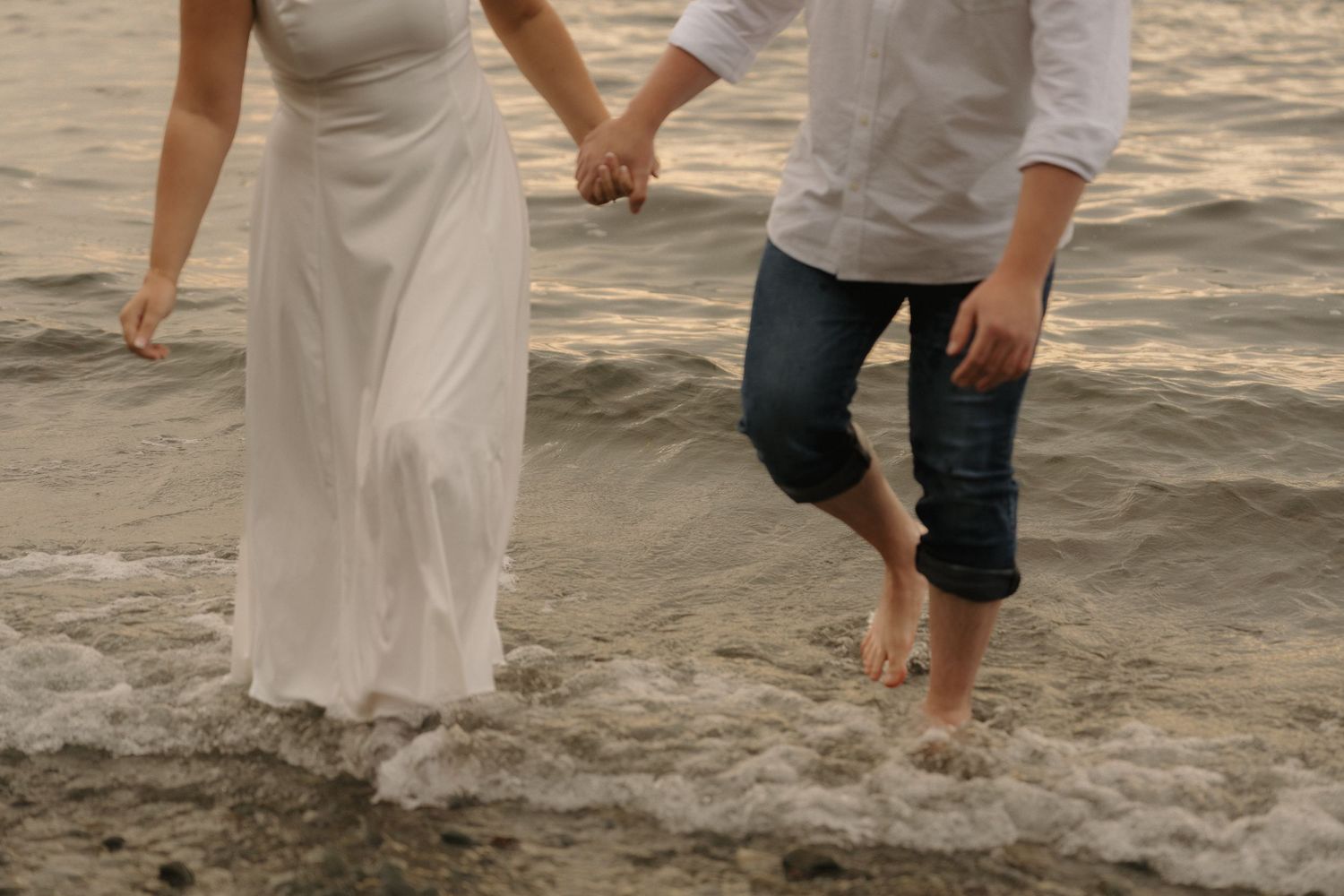 A couple holding hands and walking barefoot on a beach, with the woman wearing a flowing white dress.