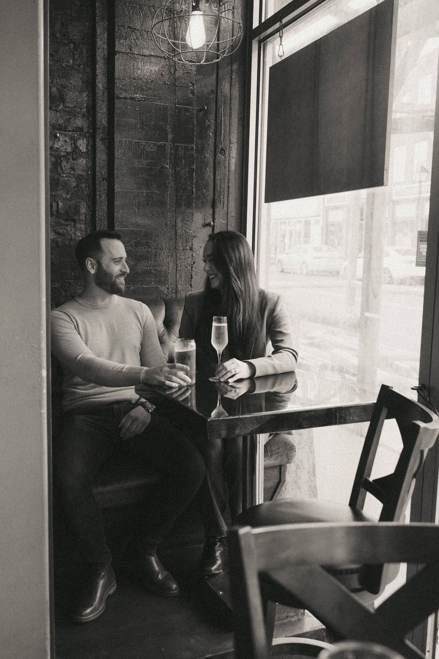 Black and white photo of a couple enjoying champagne at a cafe table by a window.
