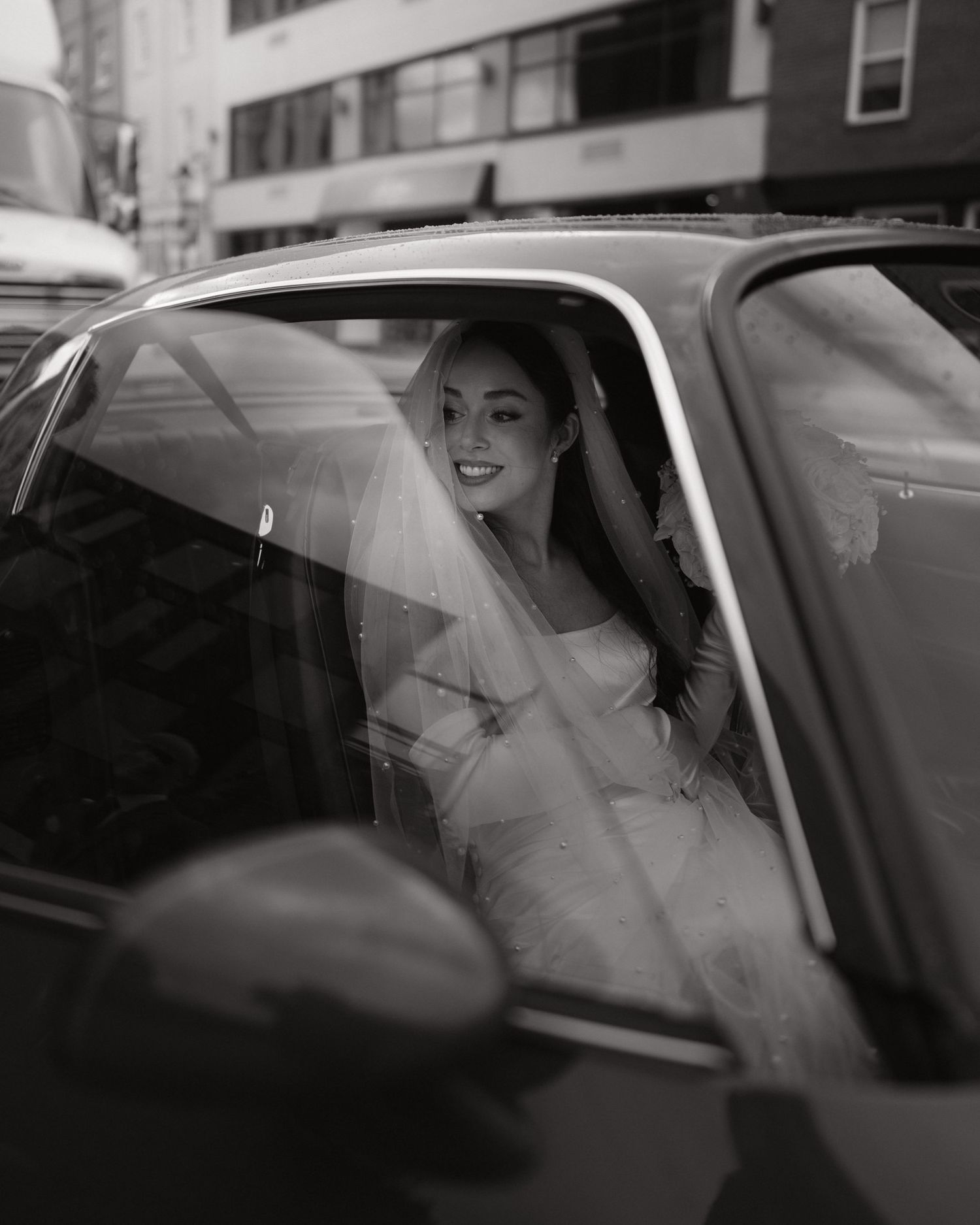 Black and white photo of a smiling bride in a wedding dress and veil sitting in a car on a city street.