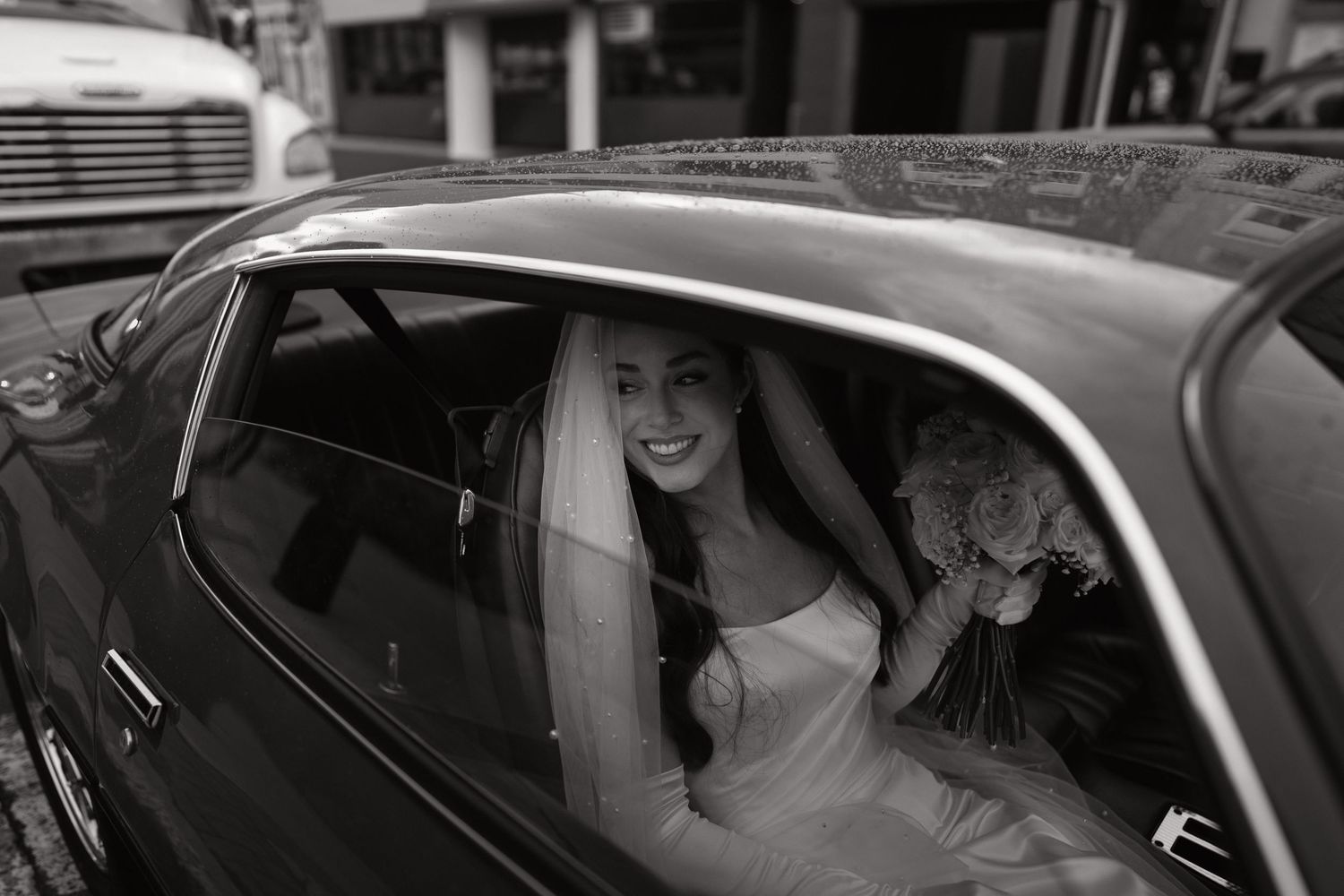 Smiling bride in wedding dress and veil sitting in a vintage car holding a bouquet, black and white photo.