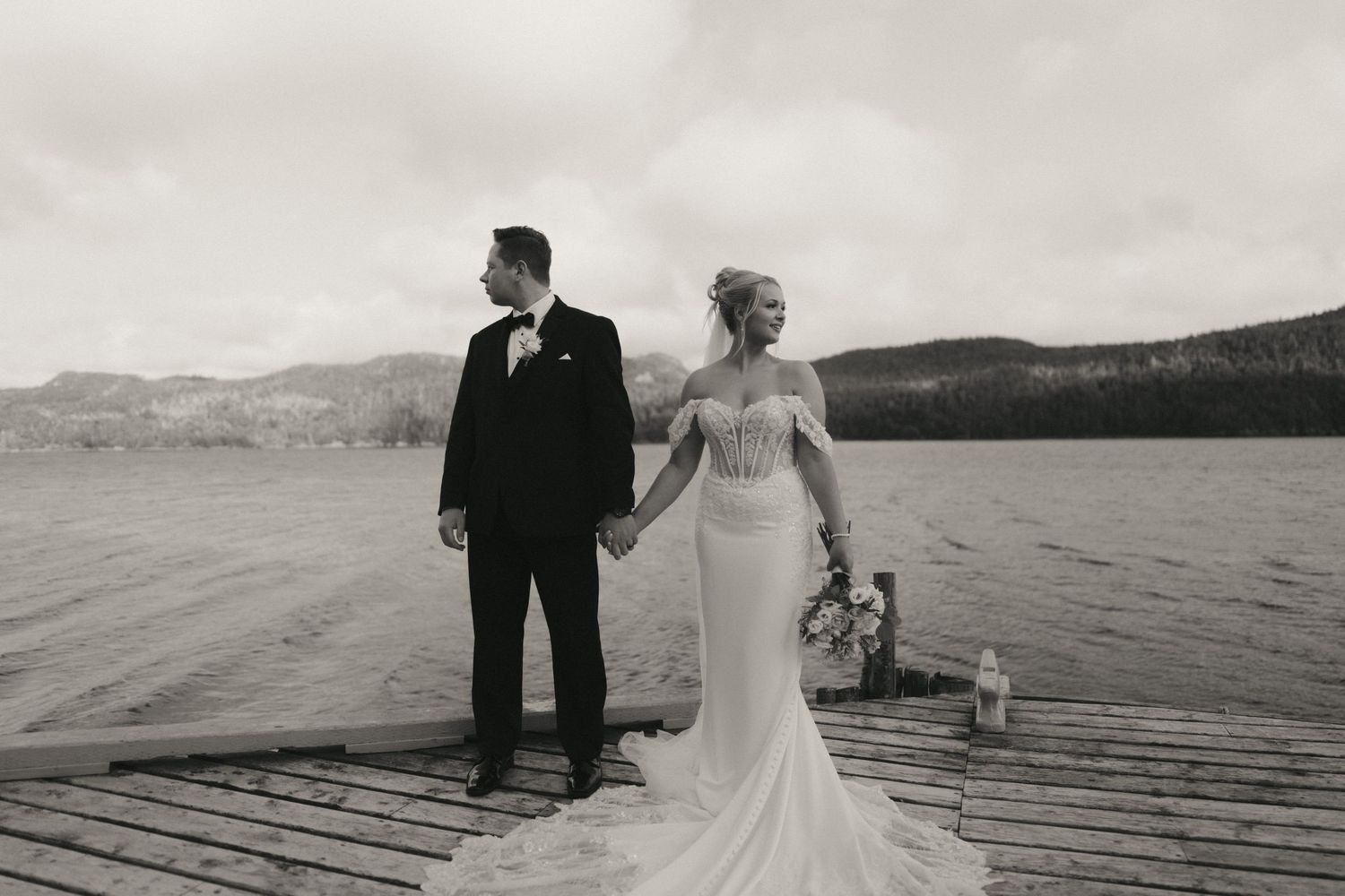 Bride and groom standing on a wooden dock by a lake in black and white wedding portrait.