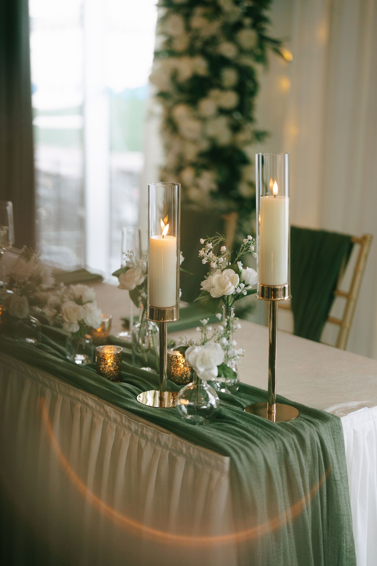 Elegant wedding table with gold candle holders, white candles, green draped runner, and white floral arrangements.