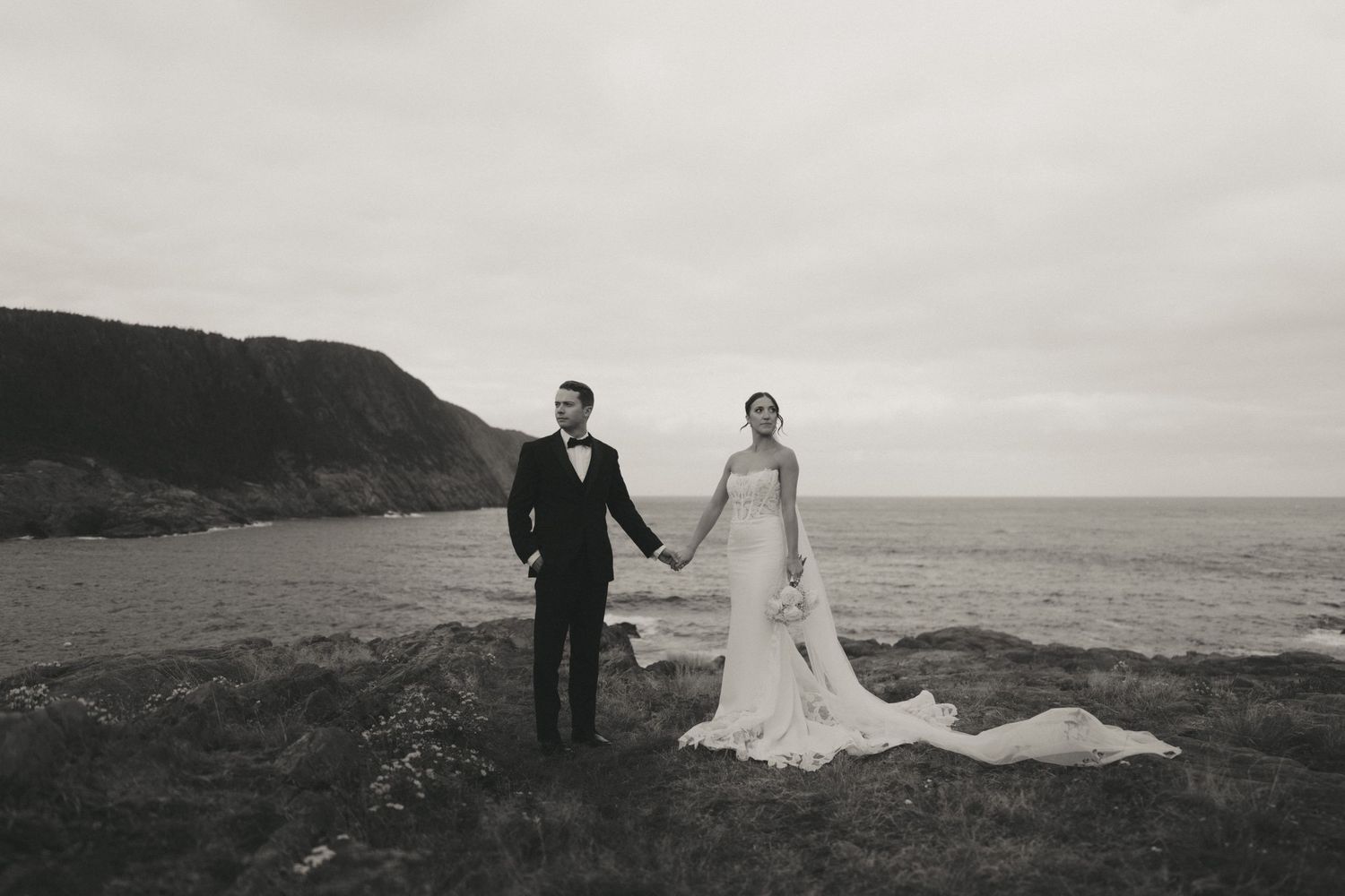 Black and white photo of a bride and groom holding hands on a coastal cliff with ocean and hills in the background.