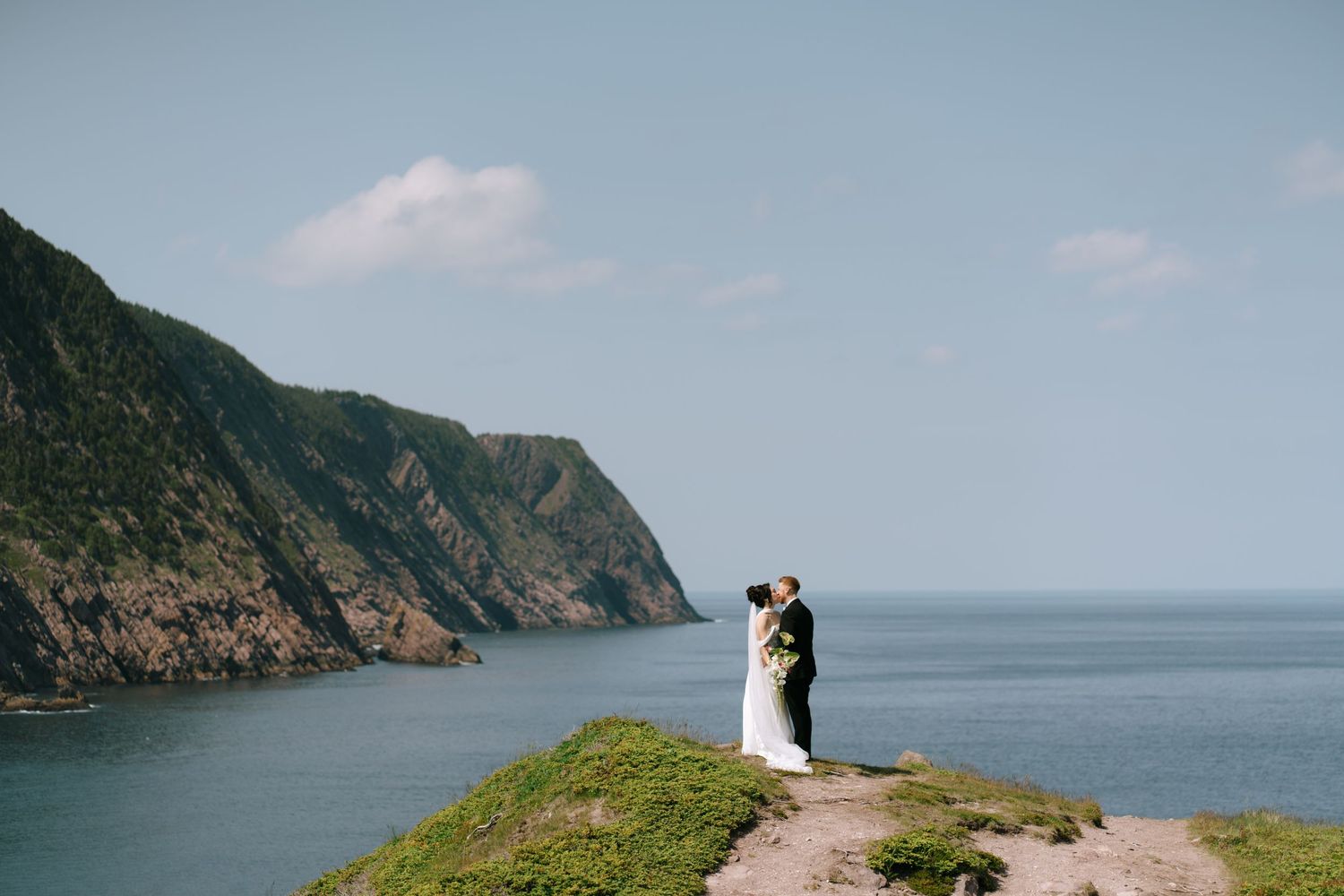 Couple sharing a romantic kiss on a coastal cliff overlooking stunning blue ocean waters and lush green mountains.