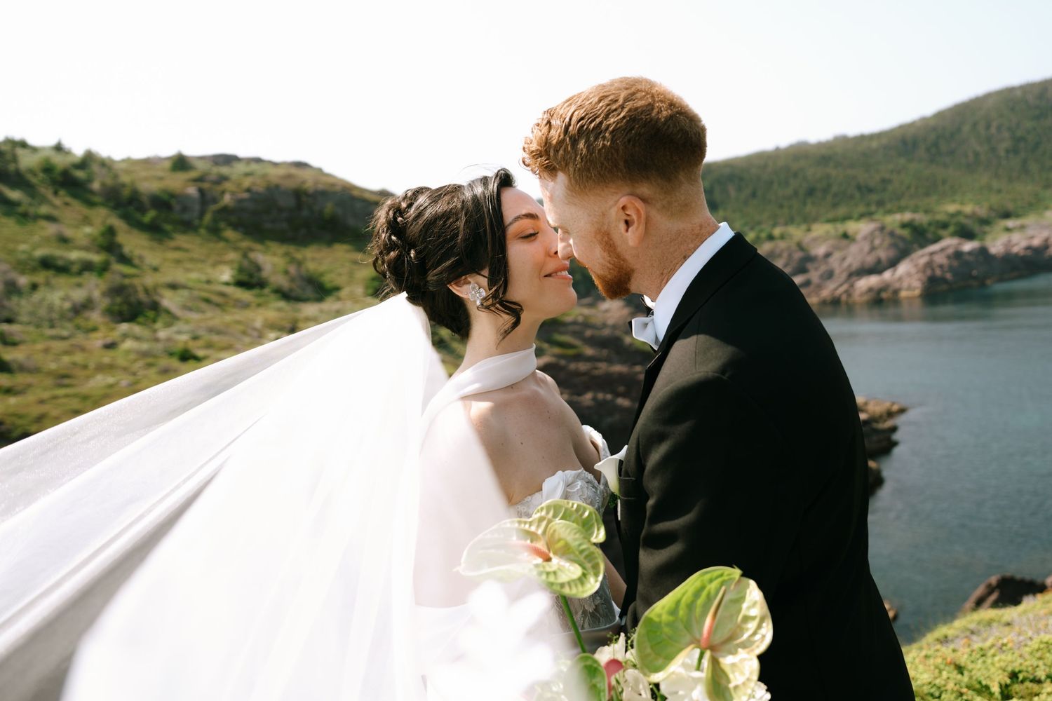 A bride and groom share a romantic kiss outdoors with scenic green hills and ocean cliffs in the background.
