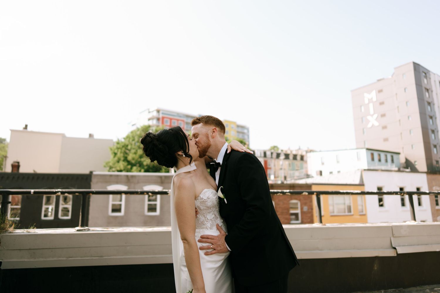 A bride and groom share a romantic rooftop kiss in an urban setting, with city buildings visible in the background.