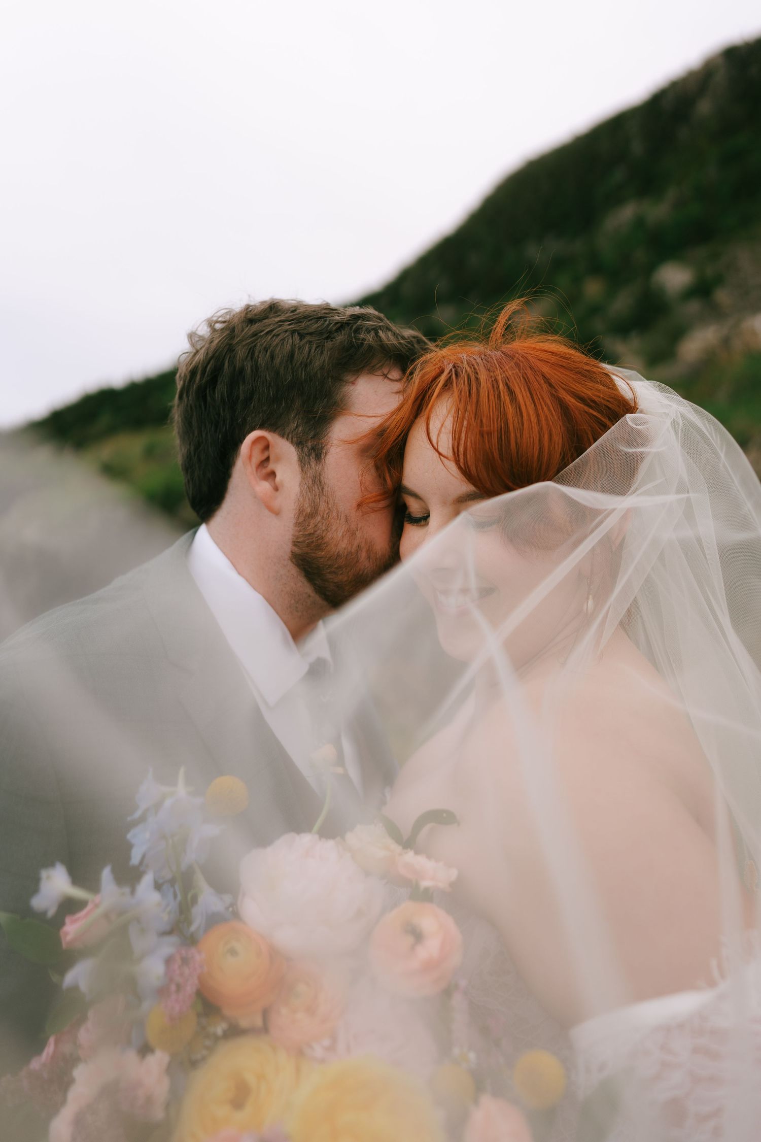 A bride and groom share a tender kiss under a flowing veil, surrounded by a colorful bouquet with a scenic hillside backdrop.
