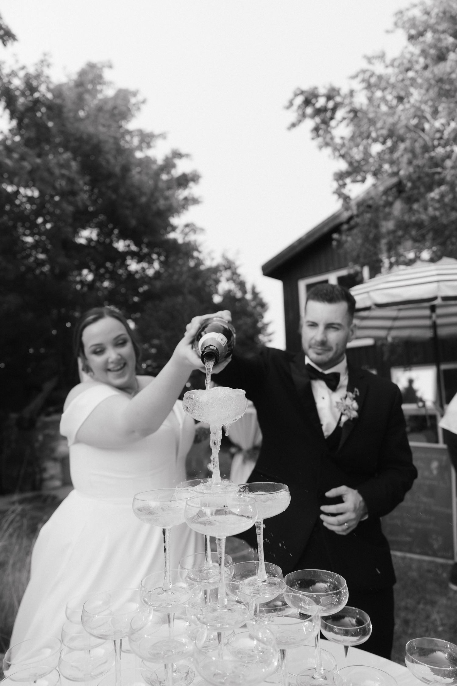 Black and white photo of wedding couple pouring champagne into a crystal glass tower outdoors.